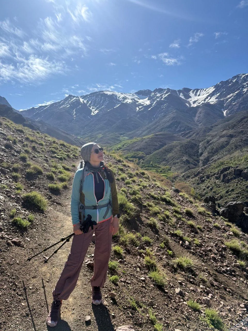 Trekker on a green High Atlas valley trail with snow-capped peaks behind